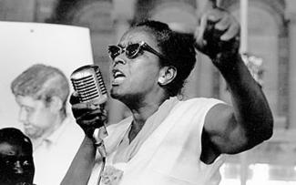 Ella Baker speaking at a microphone with her left hand raised.