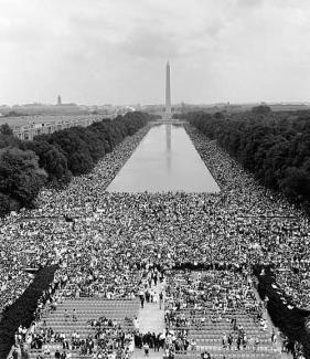 Black and white photo from high on the Lincoln Memorial looking down at crowds of people covering the National Mall around the reflecting pool, with the Washington Monument in the background.