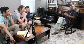 Students seated on couch at left, video camera center, Dr. Leslie Burl McLemore seated in rocking chair in front of fireplace at right