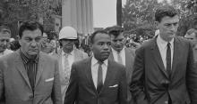 James Meredith, a black man, flanked by white U.S. Marshalls, followed by a crowd as he walks to class on the campus of Mississippi University.