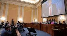 Committee members seated at a raised desk at front of room; Donald Trump photo on large screen above committee members; press photographers in foreground.