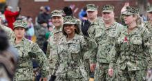 Service members walk down a street and wave to a crowd gathered on the sidewalk.