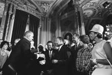 President Lyndon B. Johnson moves to shake hands with Dr. Martin Luther King Jr. with pen in hand while others look on 